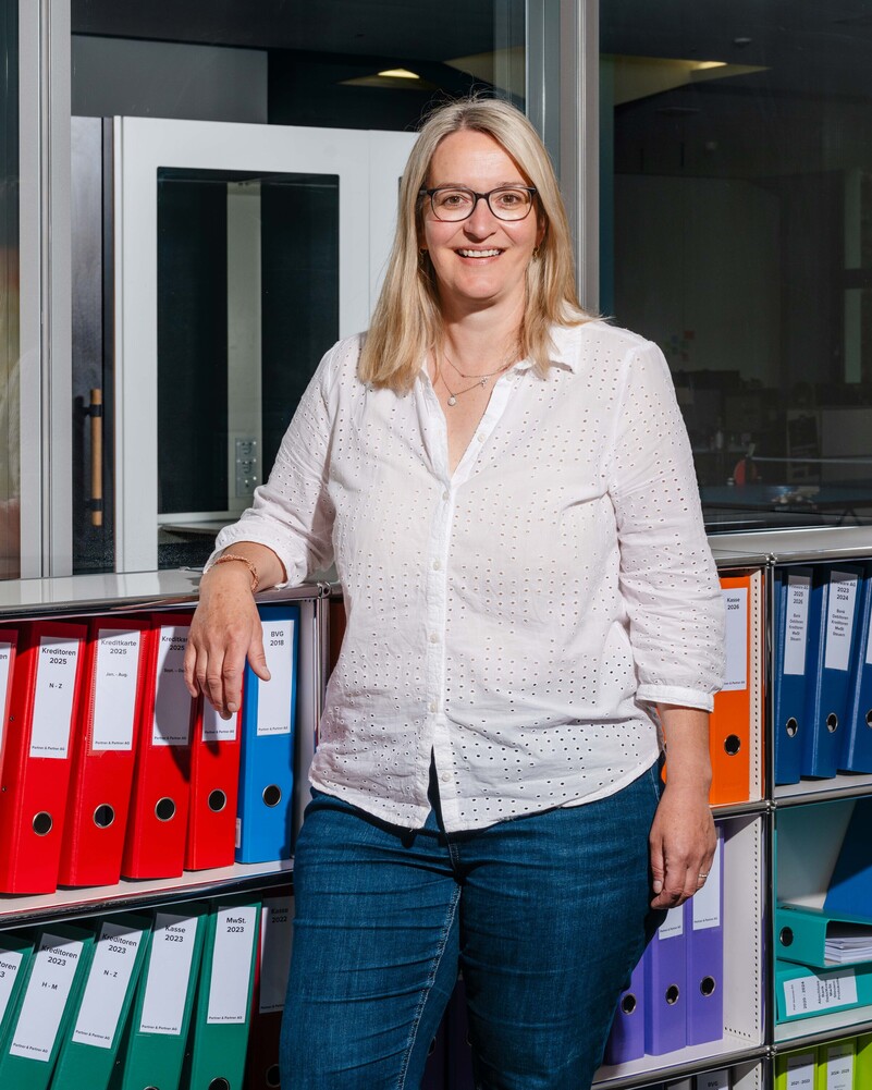 Heidi Munderich stands smiling in front of shelves filled with colourful binders.