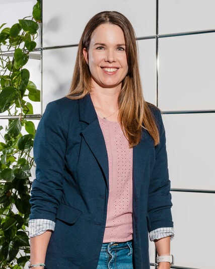 Sabine Weber stands smiling beside a plant and the company symbol.
