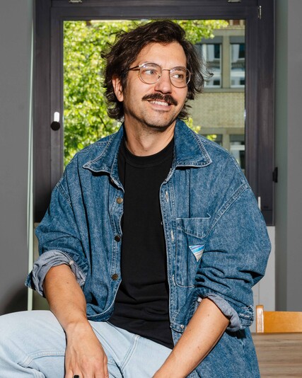 Gianfranco Acocella sits on a wooden table among chairs.
