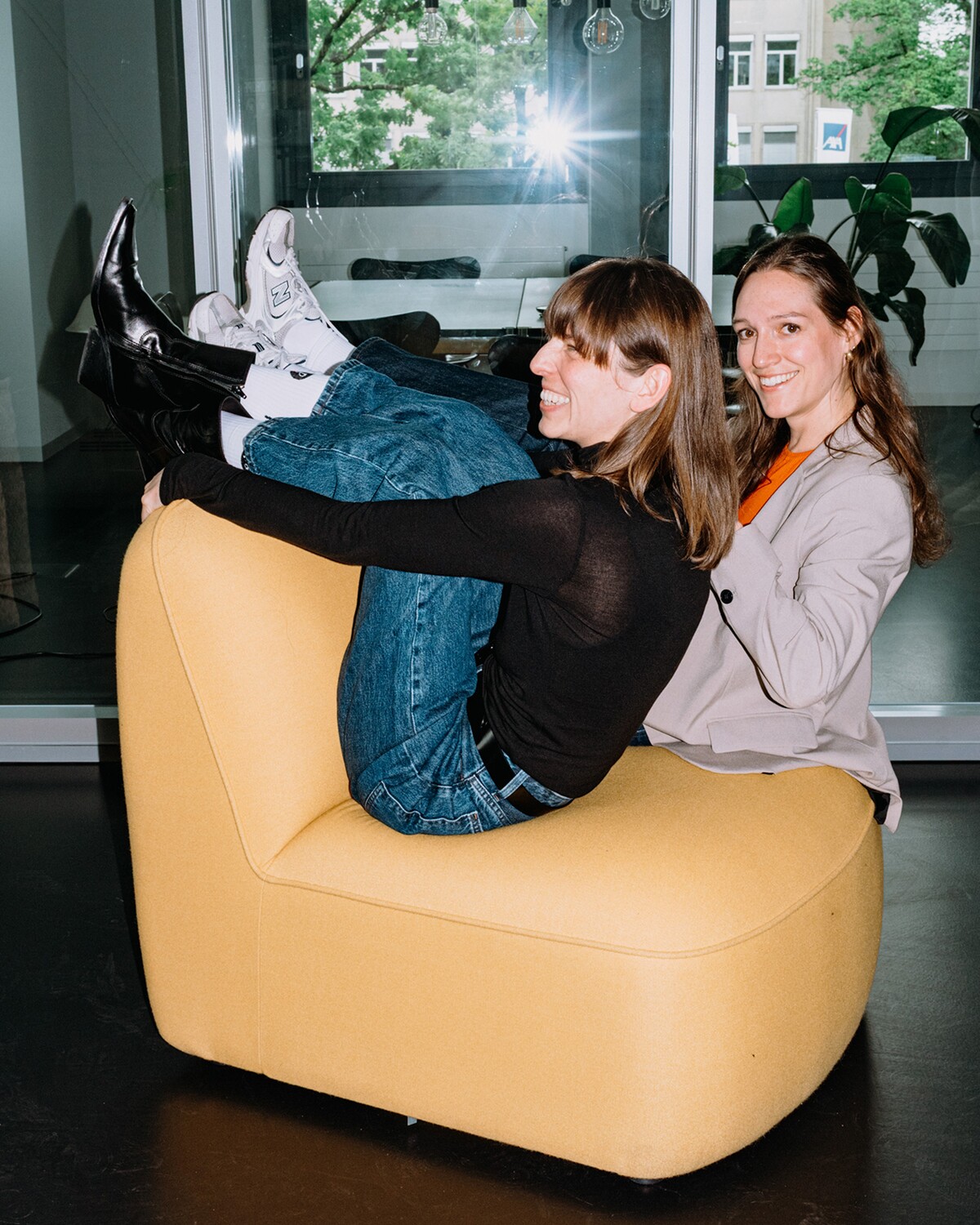 Two colleagues sit laughing together on a yellow armchair.