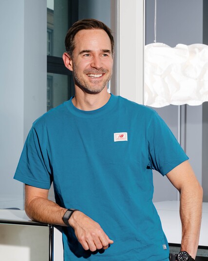 Luc Holzach leans smiling against a white shelving unit.