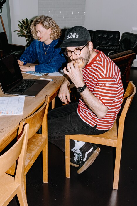 Two team members work at a table while one eats a sandwich.