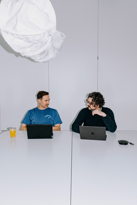 Two colleagues with laptops sit opposite each other at a long table.