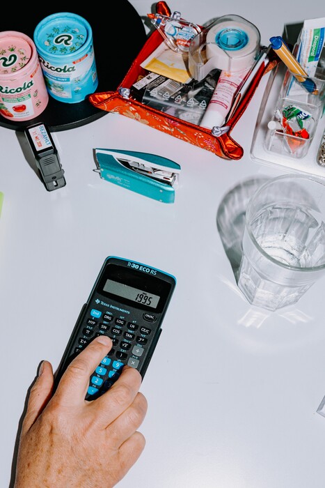 A hand uses a calculator among office and kitchen supplies.