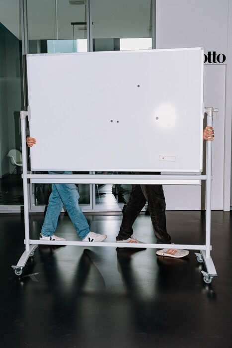 Two people carry a large mobile whiteboard through the office.