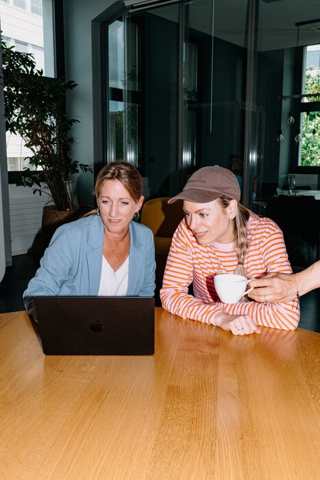 Two colleagues look at a laptop together at a wooden table.