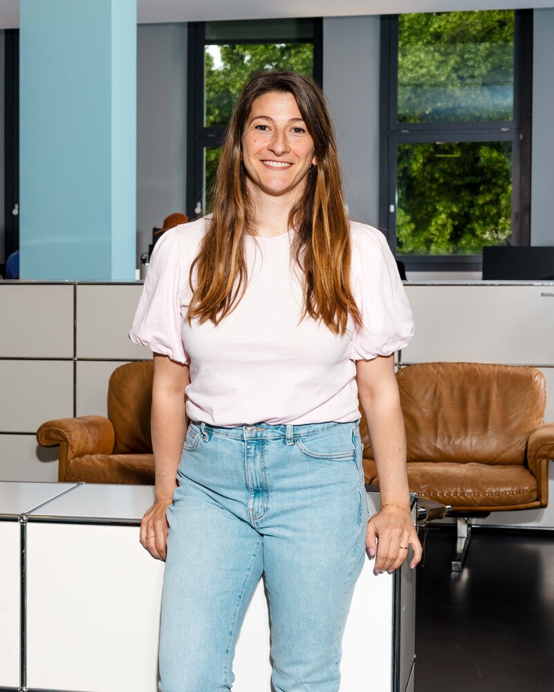 Tiziana Mascolo stands smiling in front of a brown leather sofa.