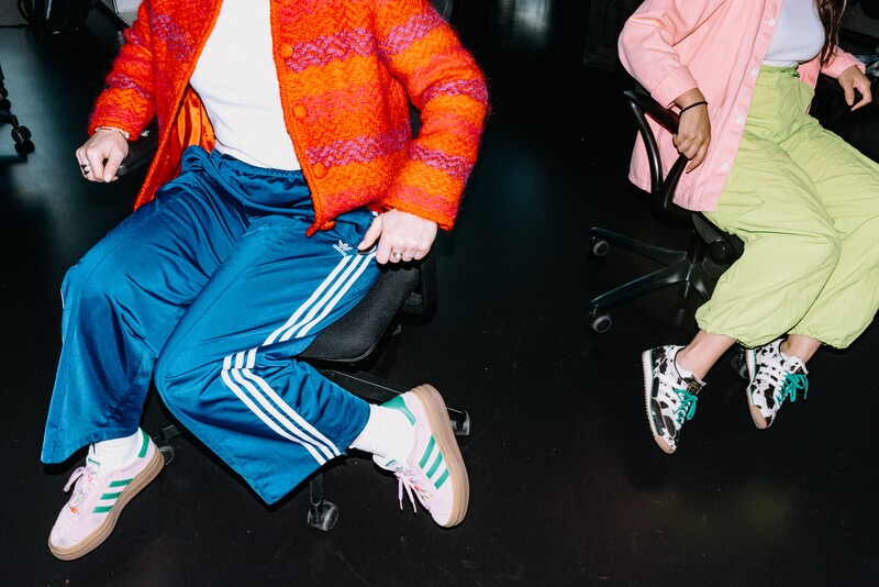 Two seated people in colourful clothes and trainers, cropped to show their legs and feet.