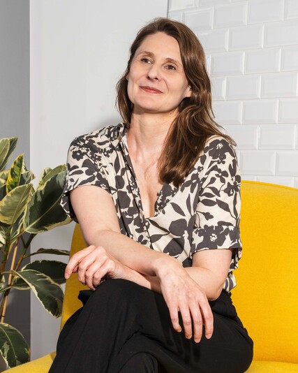 Sarah Pally sits on a yellow armchair beside a large houseplant.