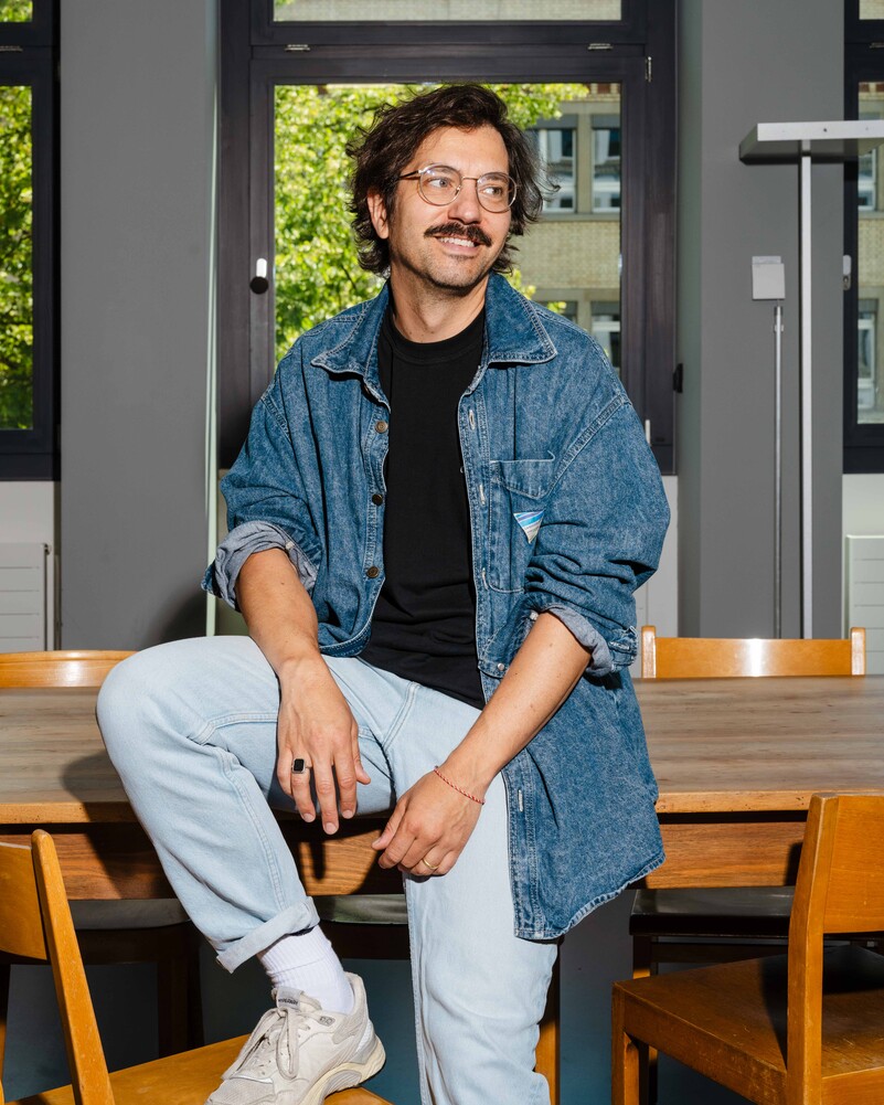 Gianfranco Acocella sits on a wooden table among chairs.