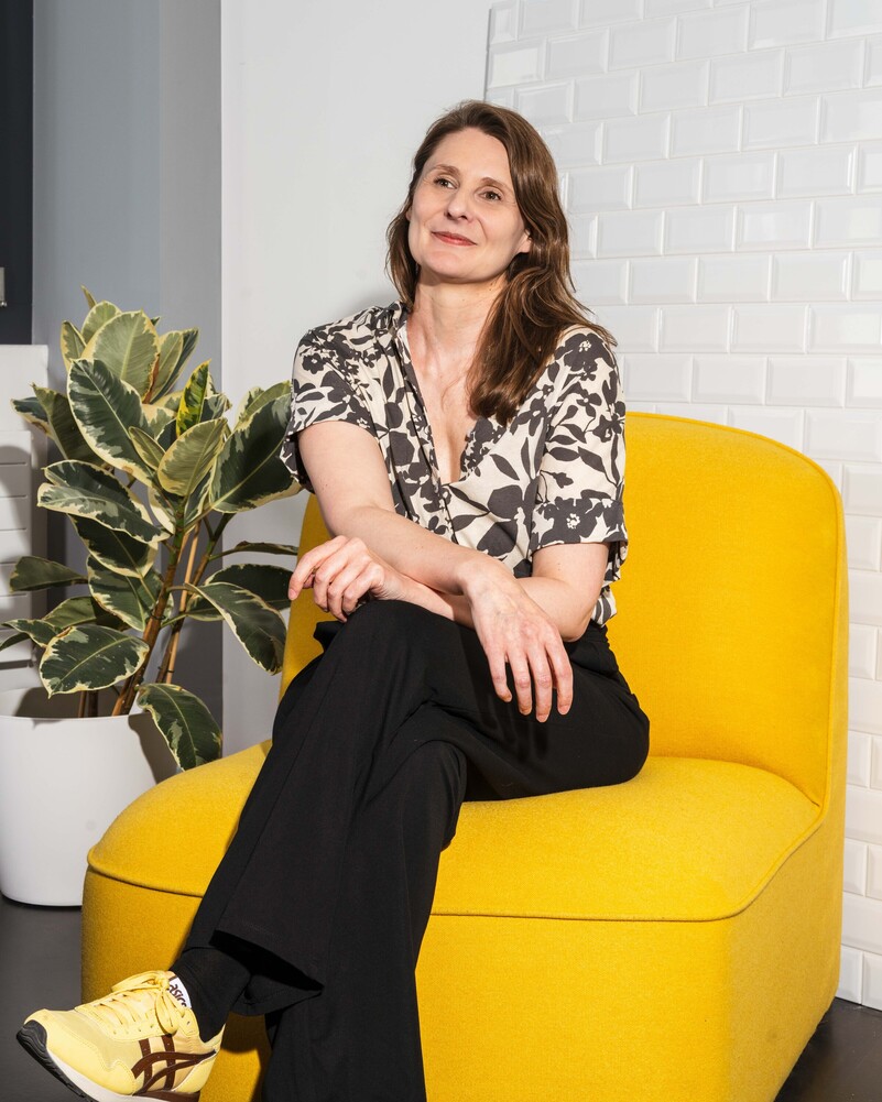 Sarah Pally sits on a yellow armchair beside a large houseplant.