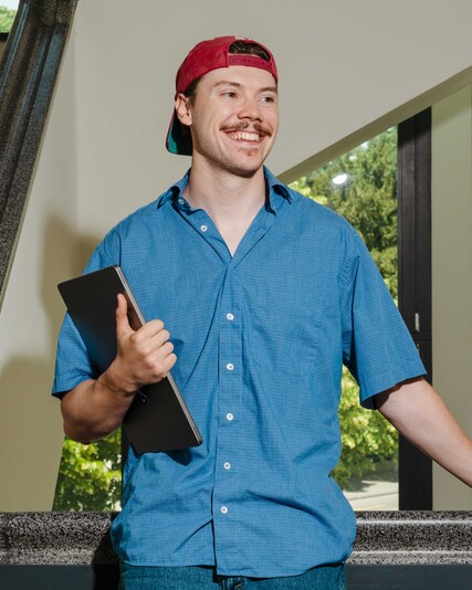 Dennis Jola stands beside a staircase holding a black folder.