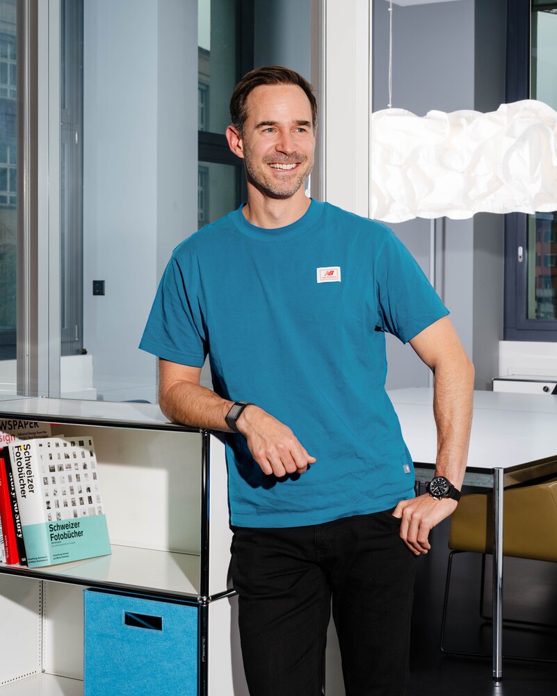 Luc Holzach leans smiling against a white shelving unit.