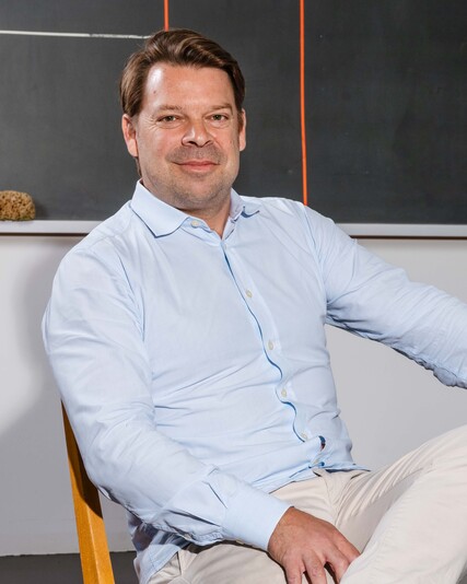 Manuel Specker sits at a wooden table with his legs crossed.