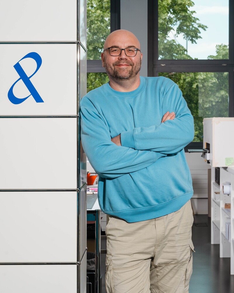 Adrian Dryja leans against a white shelving unit with his arms crossed.