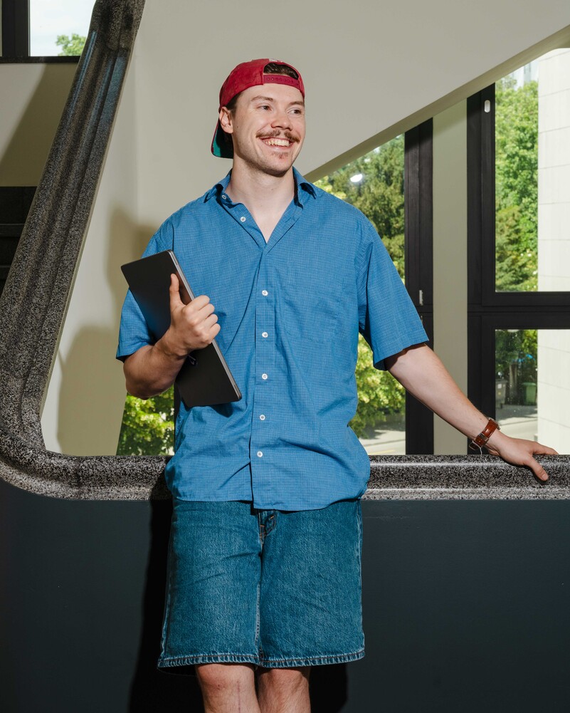 Dennis Jola stands beside a staircase holding a black folder.