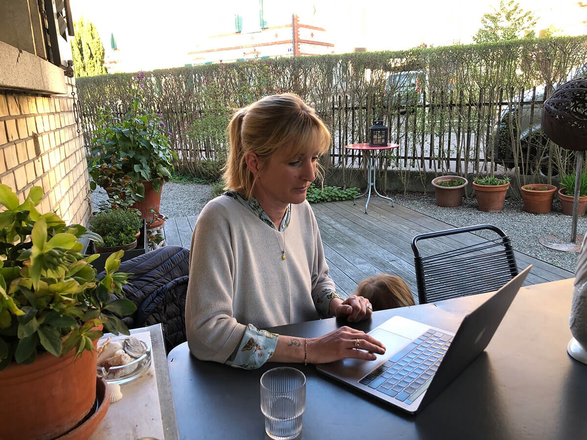 A person works on a laptop at a garden table while a child stands nearby.