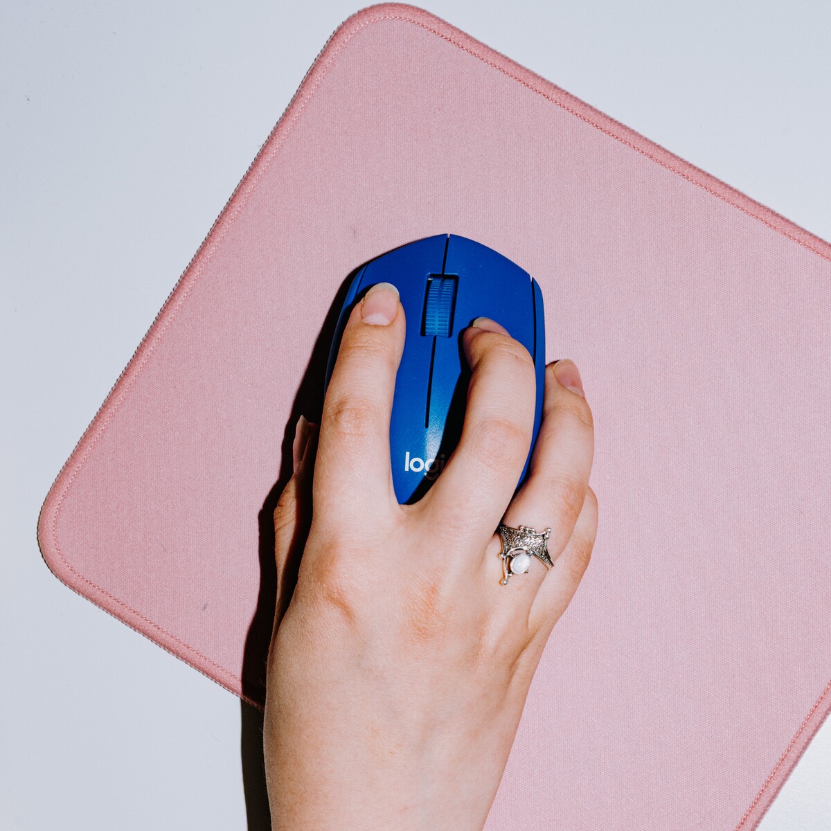 A hand operates a blue computer mouse on a pink mat.