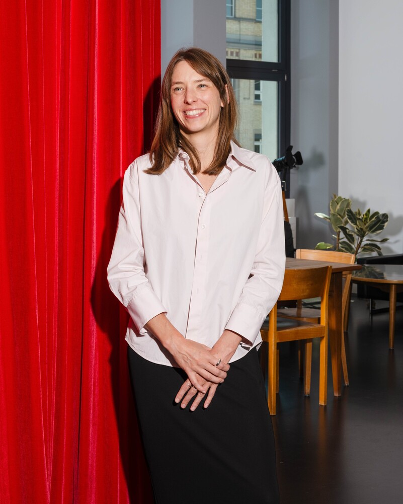Lena Thomaka stands smiling in front of a red curtain.