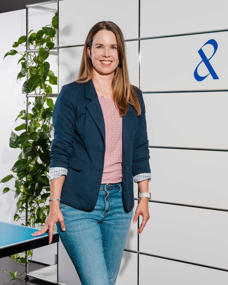 Sabine Weber stands smiling beside a plant and the company symbol.