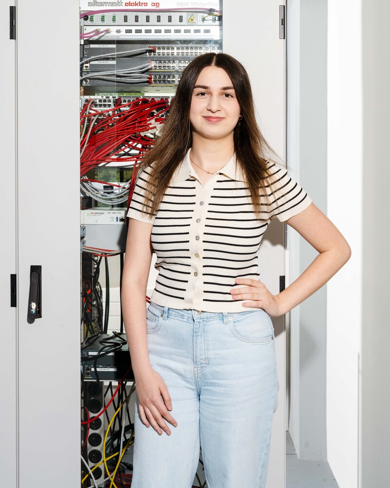 Sena Besir stands in front of an open server rack with red network cables.