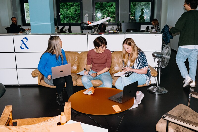 Three colleagues discuss documents and laptops on a leather sofa.