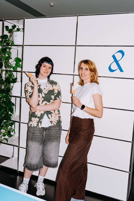Two colleagues stand with paint rollers in front of the company symbol.